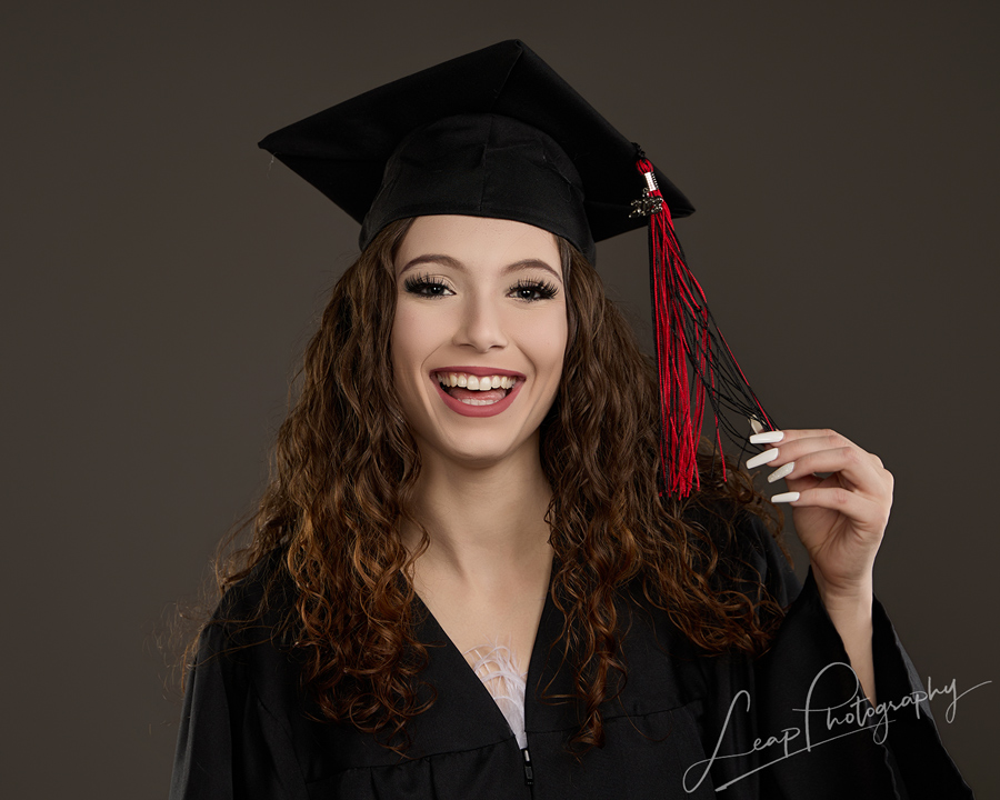 Studio headshot of a smiling high school senior wearing a black cap and gown while twirling her graduation tassel, photographed against a gray seamless background.