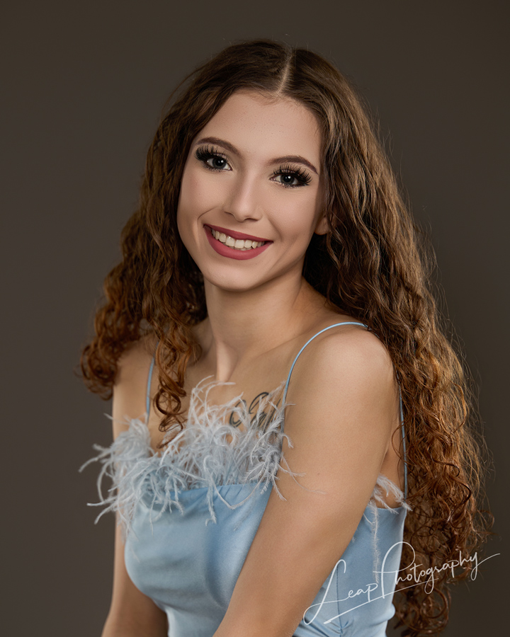 Traditional studio headshot of a high school senior wearing a blue dress with feather detailing against a gray seamless background with soft, directional lighting and subtle rim light.